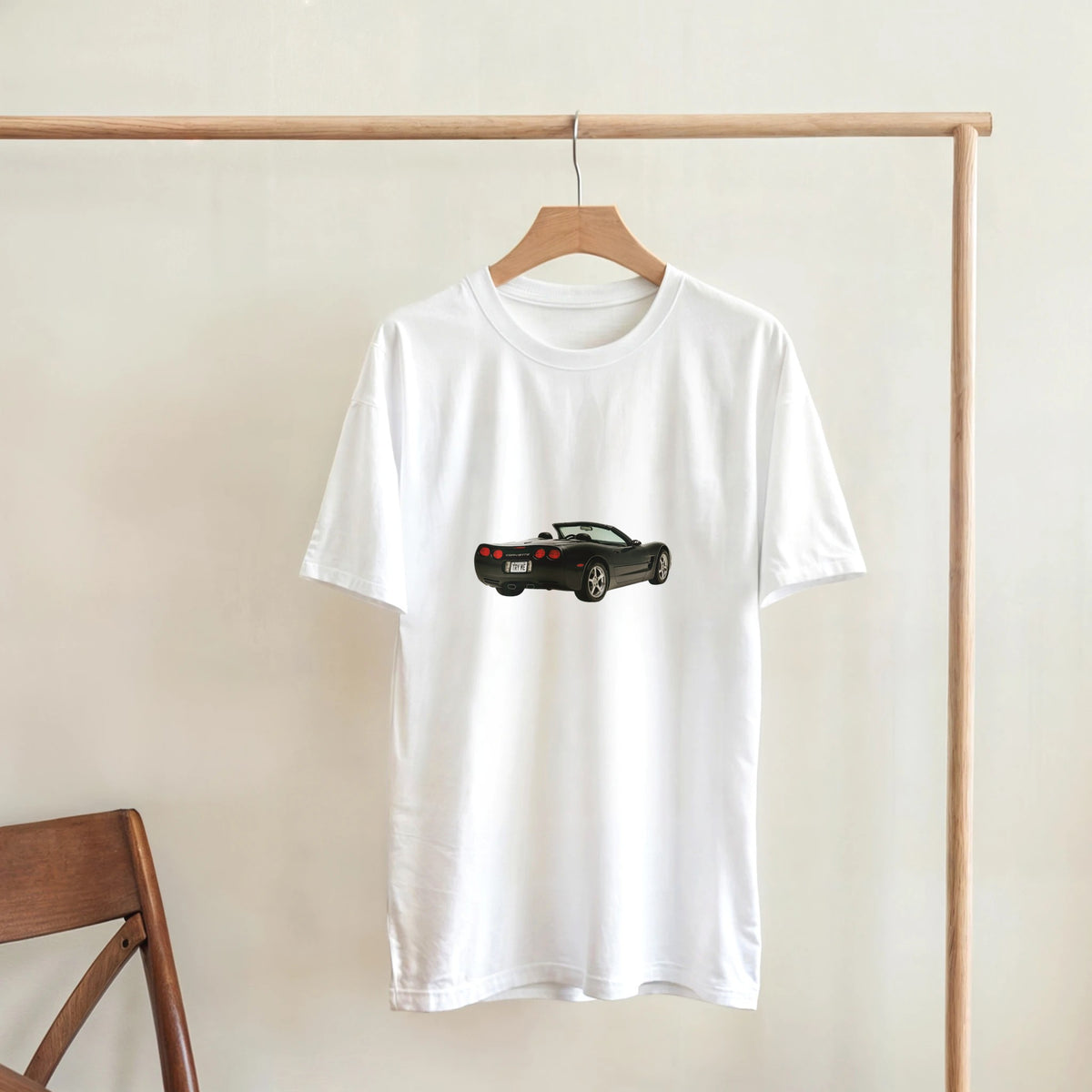 White t-shirt with a black car graphic on a wooden hanger against a beige wall.